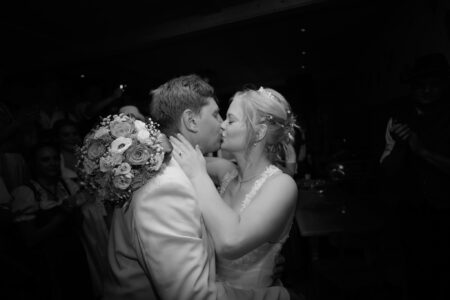 Schwarz-Weiß-Foto: Braut und Bräutigam tanzen Arm in Arm, Wange an Wange, allein angestrahlt im dunklen Saal bei der Hochzeit