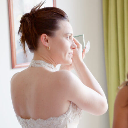 Braut trägt vor dem Fenster mit Handspiegel Lippenstift auf, vorbereitet zum Hochzeitstag