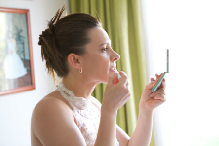 Braut trägt vor dem Fenster mit Handspiegel Lippenstift auf, vorbereitet zum Hochzeitstag