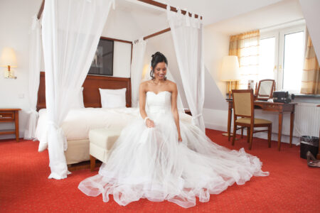 Braut sitzt im Hochzeitskleid auf rotem Teppich vor dem Bett in der Hochzeitssuite und blickt nach unten – Get Ready