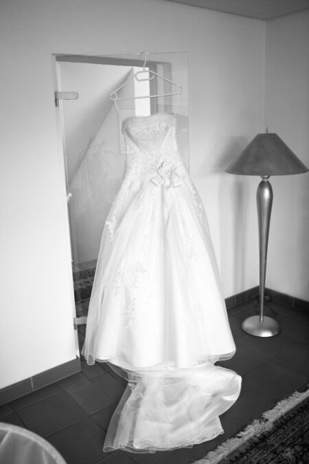Schwarz-Weiß-Foto eines Brautkleids an der Tür, daneben eine Stehlampe – Detail Hochzeitsvorbereitung